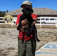 a young boy holding a black puppy in front of a fence