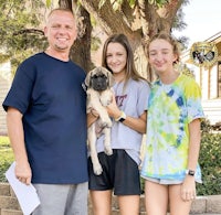 a man and two girls standing in front of a house with a pug puppy